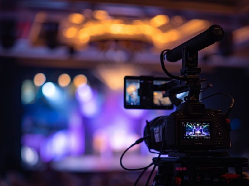 Câmera profissional de filmagem com microfone direcional captando evento corporativo em palco iluminado com painel de LED ao fundo, atmosfera dramática, fotografia realista de alto impacto